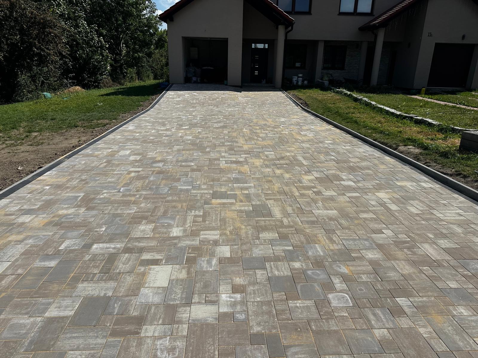 Szeroki podjazd z kostki brukowej w odcieniach szarości i beżu, obramowany szarym obrzeżem, prowadzący do domu z garażem. Widok z perspektywy osoby stojącej na podjeździe.