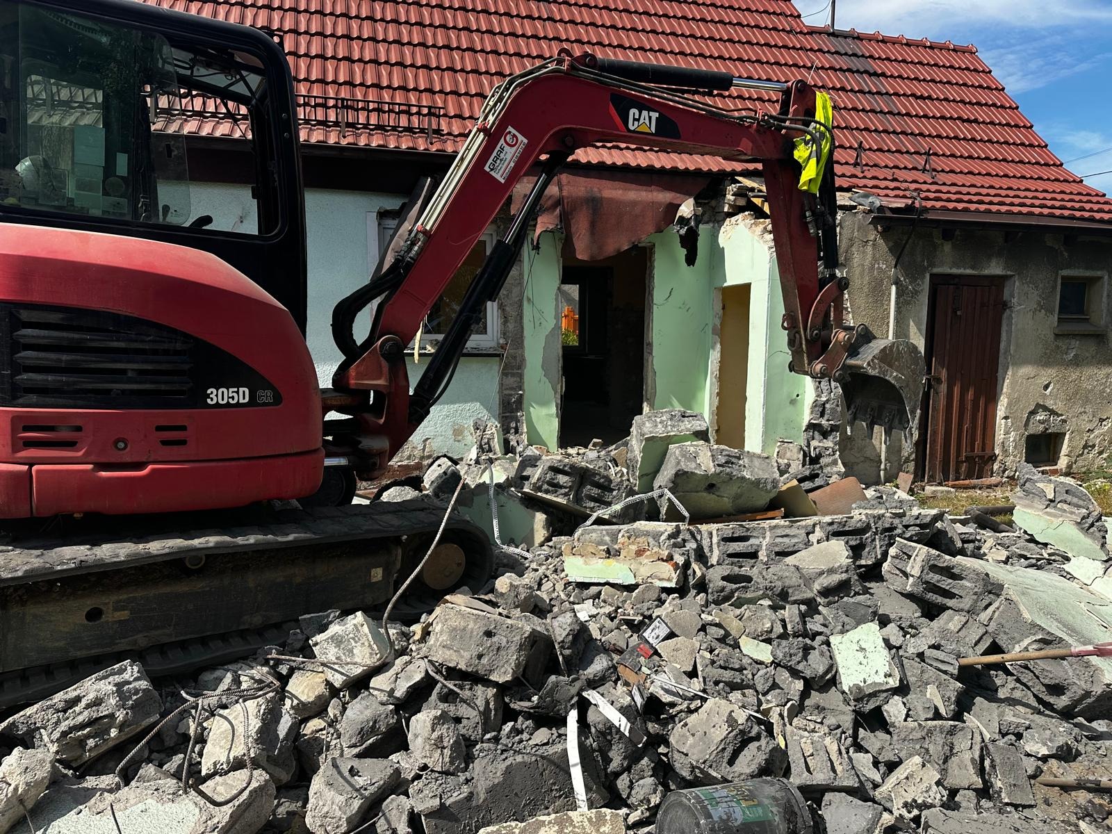 Czerwona koparka CAT 305D CR w trakcie rozbiórki starego, jednopiętrowego budynku z czerwonym dachem. Gruz i fragmenty ścian leżą na ziemi.