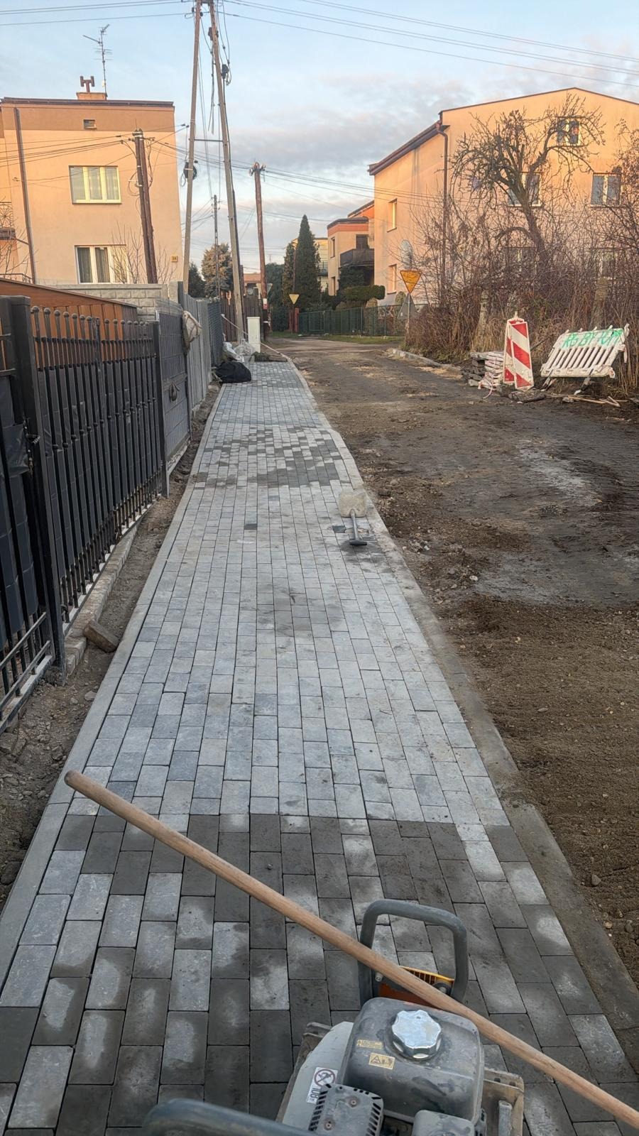 Fragment chodnika z szarej kostki brukowej w trakcie układania, widoczna ubijarka i drewniany trzonek. Częściowo ułożony chodnik sąsiaduje z ziemią i metalowym ogrodzeniem.