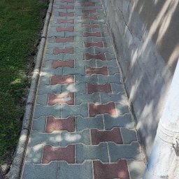 KostkaBezPodjazdu - Świeżo ułożona, dwukolorowa kostka brukowa w odcieniach szarości i czerwieni, z widocznymi resztkami piasku na powierzchni spoin, przylegająca do szarej ściany budynku.