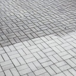 KostkaBezPodjazdu - Częściowo umyta kostka brukowa, widoczna różnica w kolorze, z urządzeniem wysokociśnieniowym w tle. Układ w jodełkę.