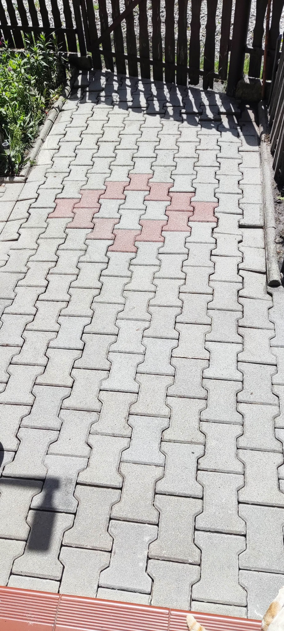 Ułożona kostka brukowa w odcieniach szarości i czerwieni, widziana z góry, z fragmentem drewnianego płotu w tle i roślinnością po lewej stronie.