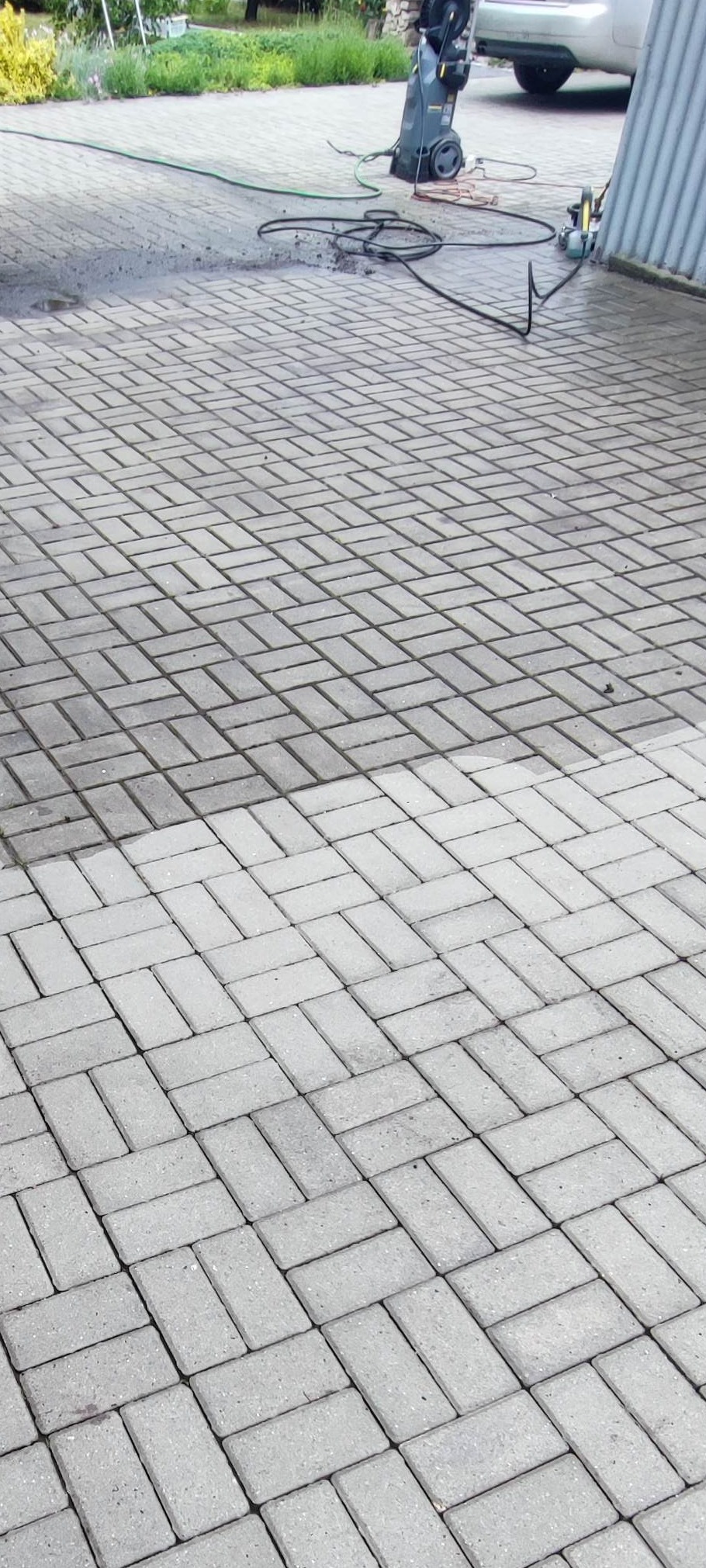 Częściowo umyta kostka brukowa, widoczna różnica w kolorze, z urządzeniem wysokociśnieniowym w tle. Układ w jodełkę.