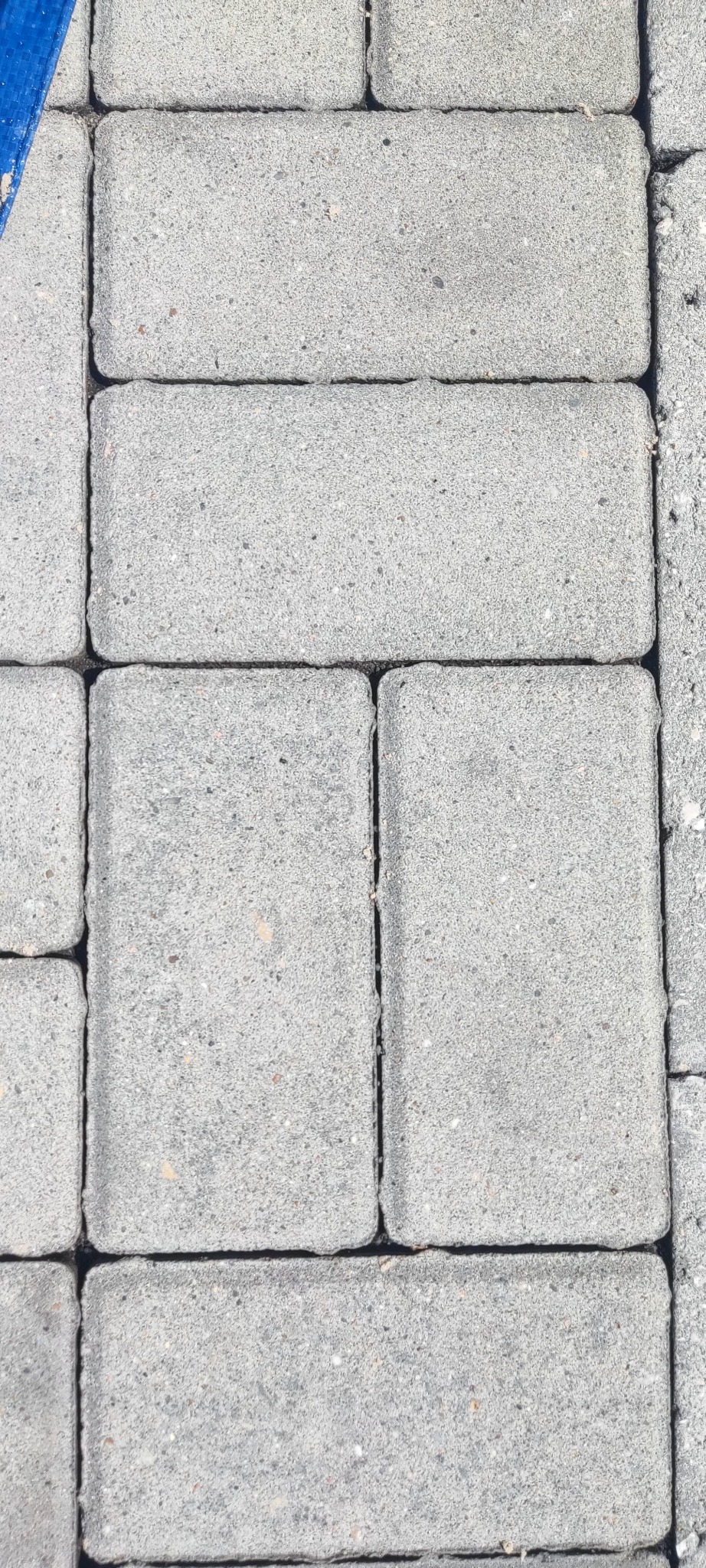 Szary wzór kostki brukowej ułożonej w regularny sposób, z widocznym fragmentem niebieskiego materiału w lewym górnym rogu.