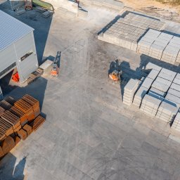 Marcin Czarnowski Firma Usługowo-Handlowa - Widok z góry na plac z prefabrykatami betonowymi, wózki widłowe i budynek magazynowy z metalową konstrukcją. Ułożone płyty tworzą regularne wzory.