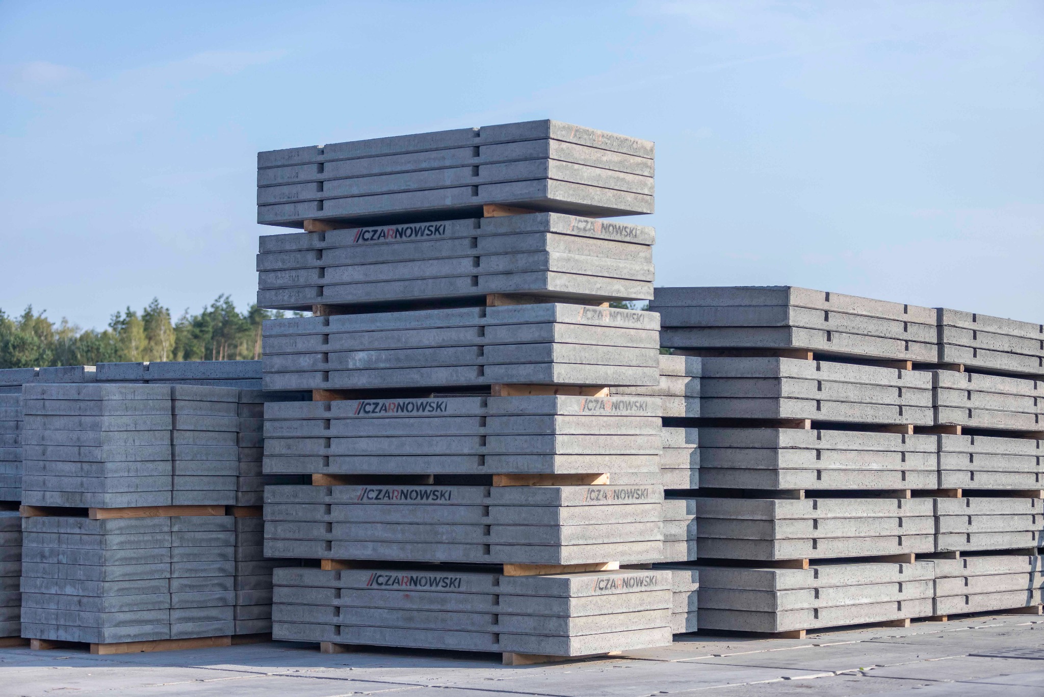 Szare płyty drogowe ułożone w stosy na paletach, widoczne napisy '//CZARNOWSKI' na płytach. Tło stanowi błękitne niebo i zieleń drzew.