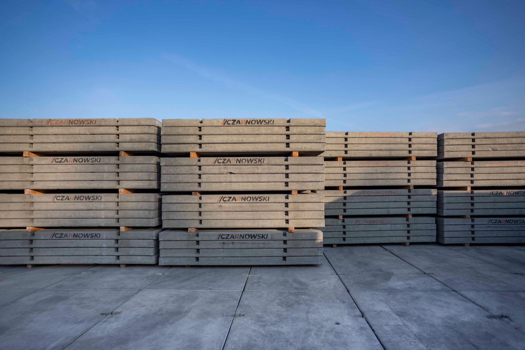 Stos prefabrykowanych, szarych płyt betonowych z logo producenta ułożonych na placu pod błękitnym niebem. Widoczne drewniane przekładki.