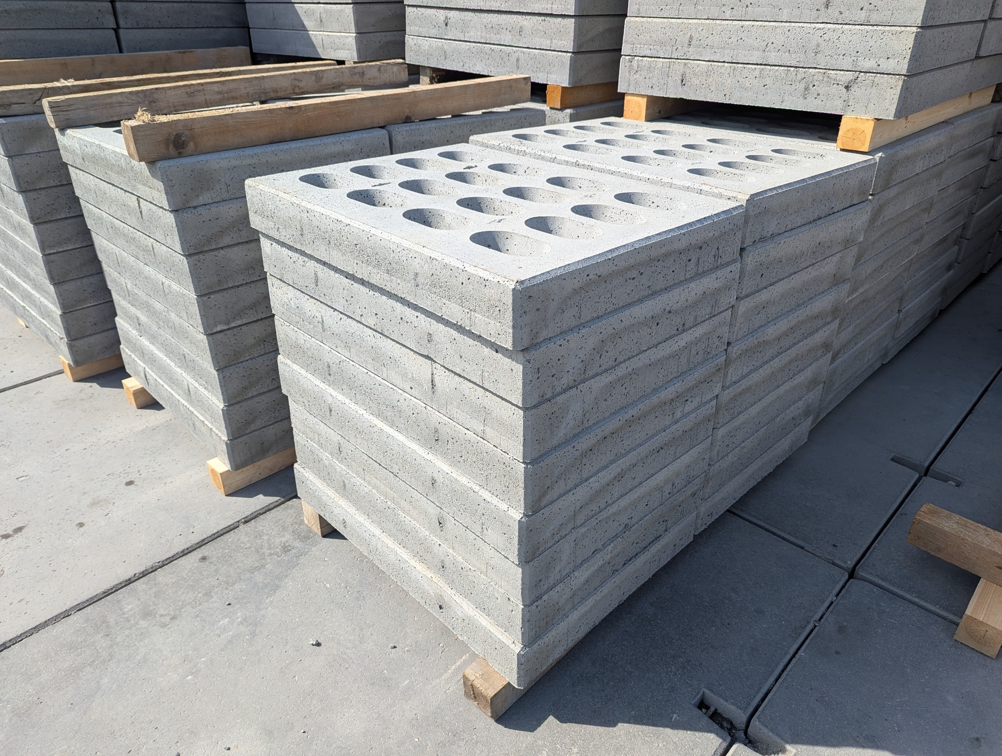 Stosy szarych, betonowych płyt drogowych z okrągłymi otworami, ułożone na drewnianych legarach. Widoczne liczne warstwy płyt i tekstura betonu.