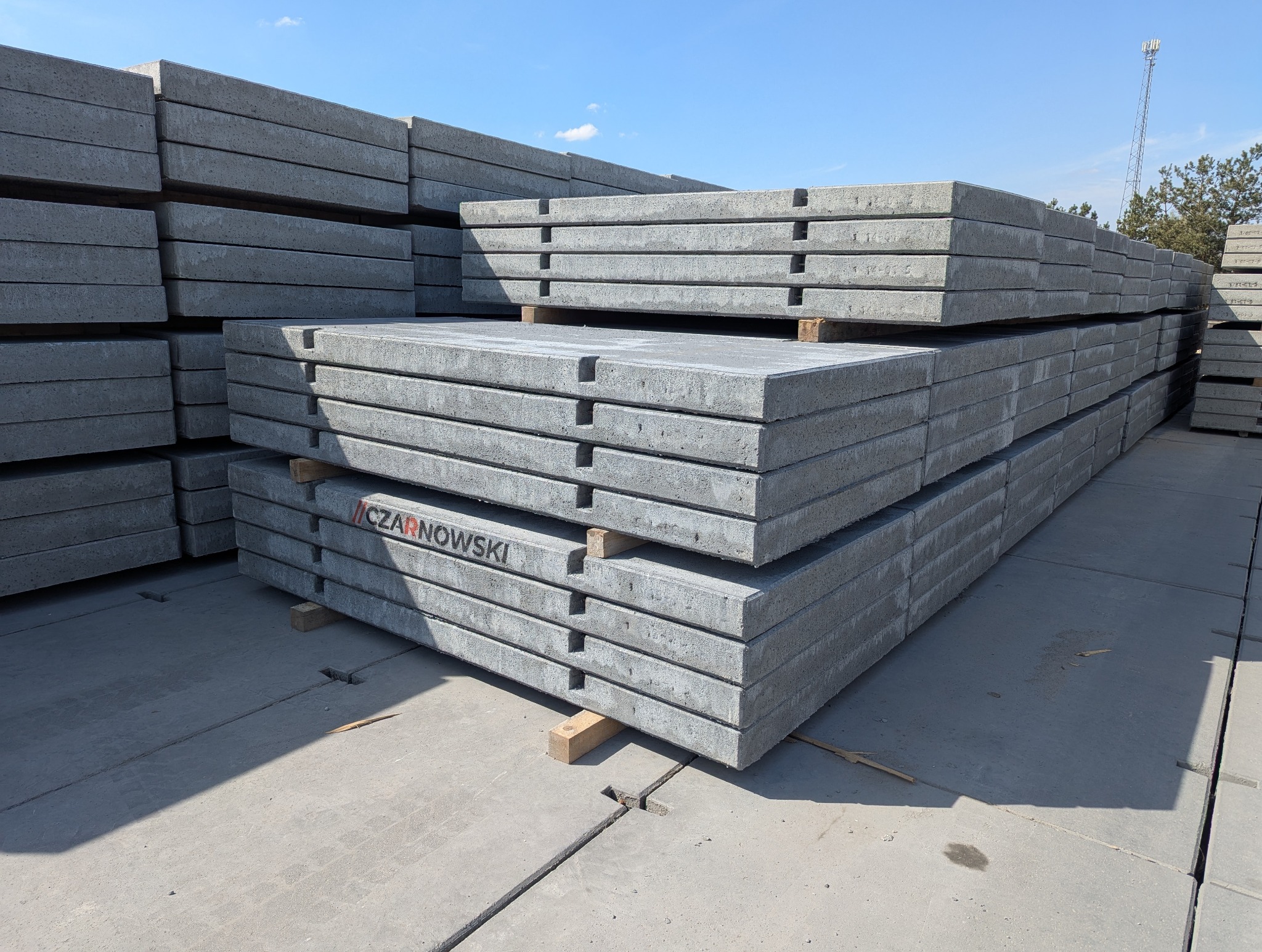 Szare płyty drogowe ułożone w stosy na placu, widoczne charakterystyczne wycięcia i oznaczenie producenta 'CZARNOWSKI'. W tle maszt telekomunikacyjny.