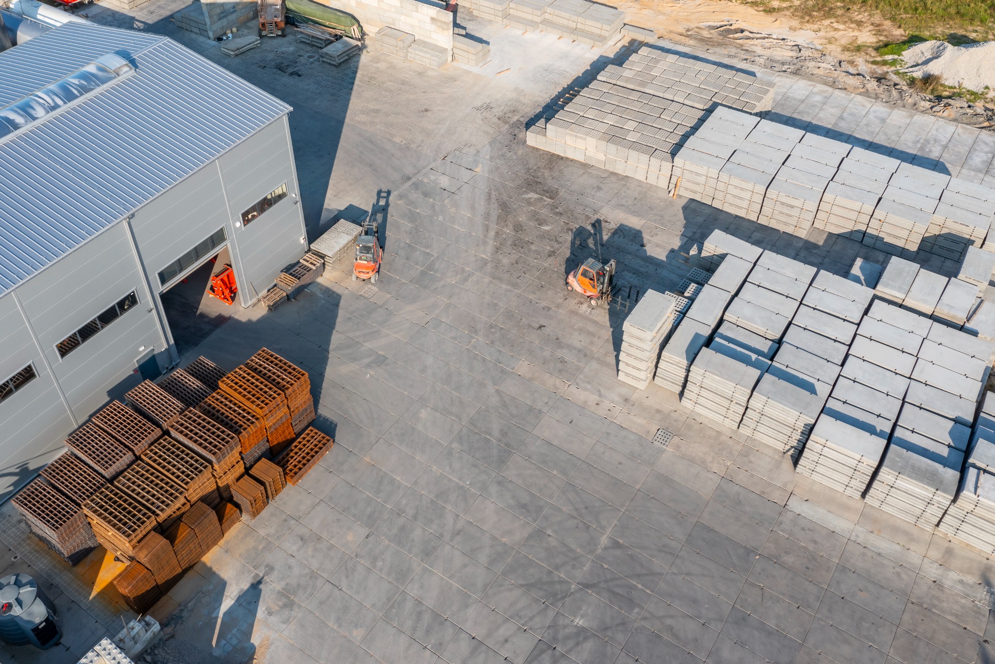 Widok z góry na plac z prefabrykatami betonowymi, wózki widłowe i budynek magazynowy z metalową konstrukcją. Ułożone płyty tworzą regularne wzory.