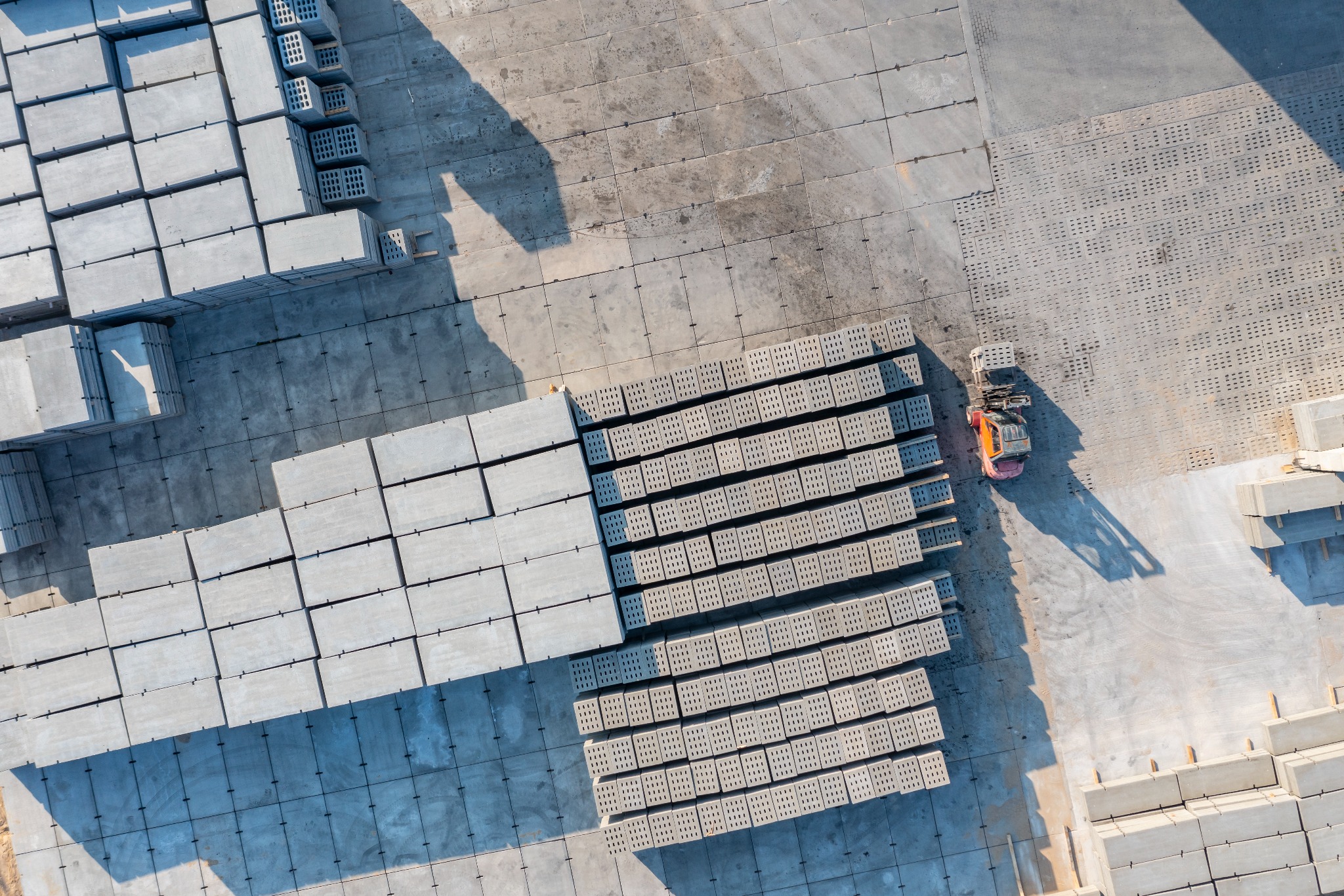 Widok z góry na plac składowy z ułożonymi prefabrykatami betonowymi i wózkiem widłowym. Silne cienie podkreślają strukturę i wzór ułożenia elementów.