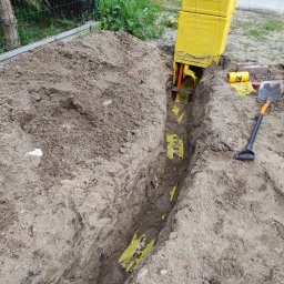 PRODUS spółka jawna K.Cygan, Z.Czuń, S.Filipiak.. - Wykop z żółtymi rurami w folii, narzędzia obok. Widok z góry na wykopaną ziemię z rurami w żółtej folii. Obok leży łopata i żółta skrzynka. Prace ziemne.