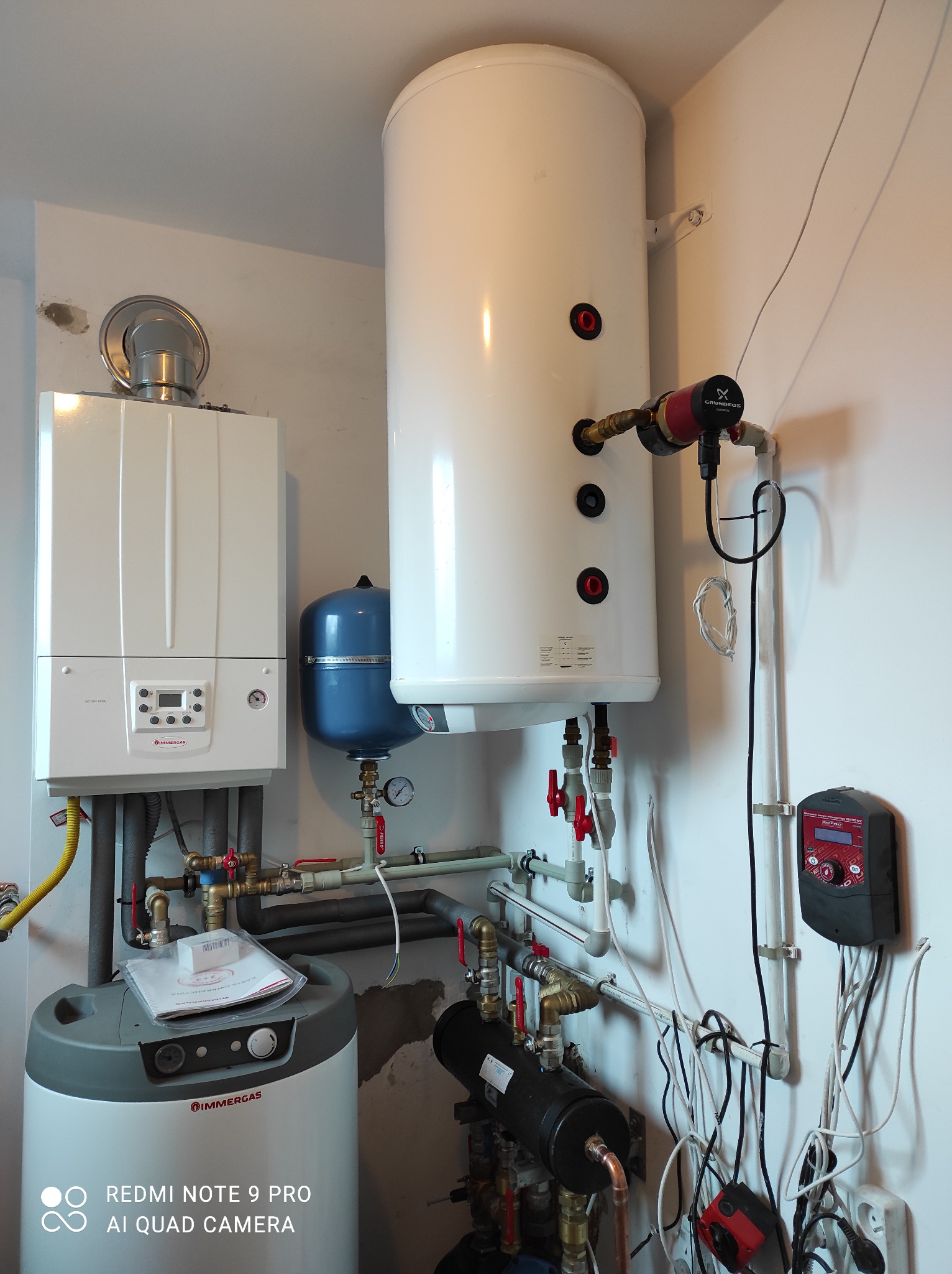 Biały piec Immergas z szarym zbiornikiem, niebieskim naczyniem wzbiorczym i białym zasobnikiem ciepłej wody, uzupełnione instalacją, widok z bliska.