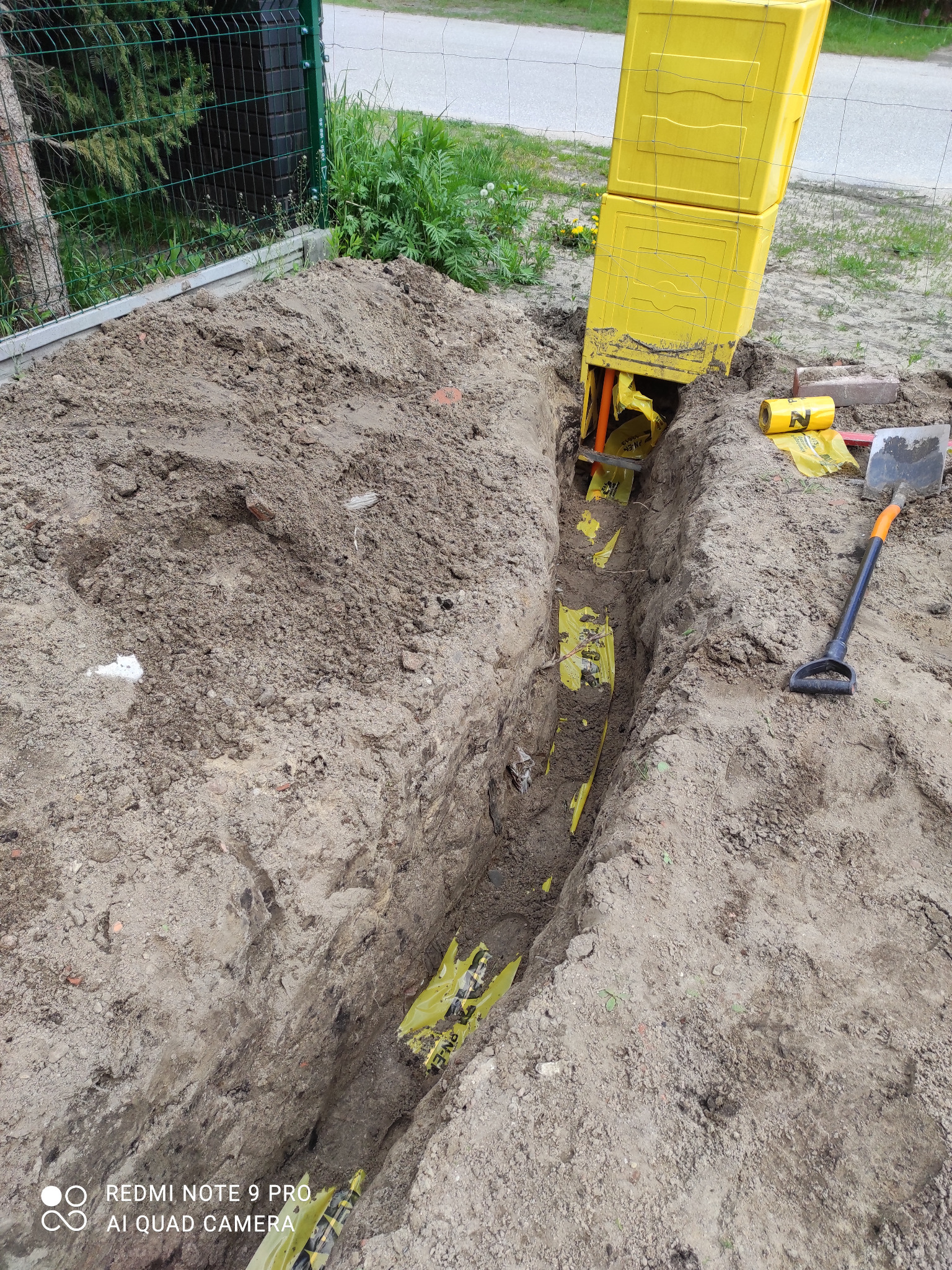 Wykop z żółtymi rurami w folii, narzędzia obok. Widok z góry na wykopaną ziemię z rurami w żółtej folii. Obok leży łopata i żółta skrzynka. Prace ziemne.