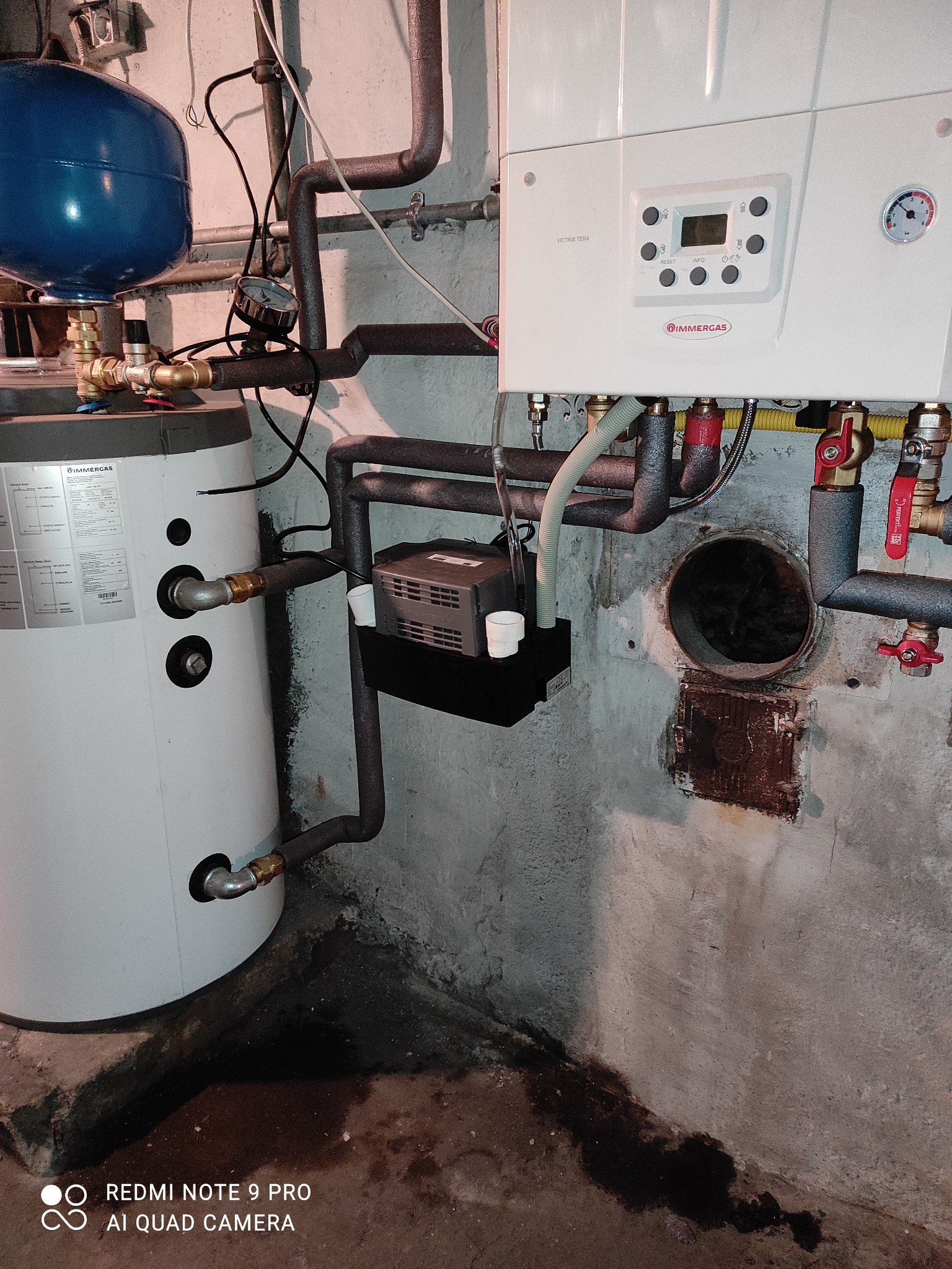 Biały piec gazowy Immergas Victrix Tera z czarnymi rurami i niebieskim zbiornikiem wyrównawczym na tle szarej, surowej ściany w piwnicy.