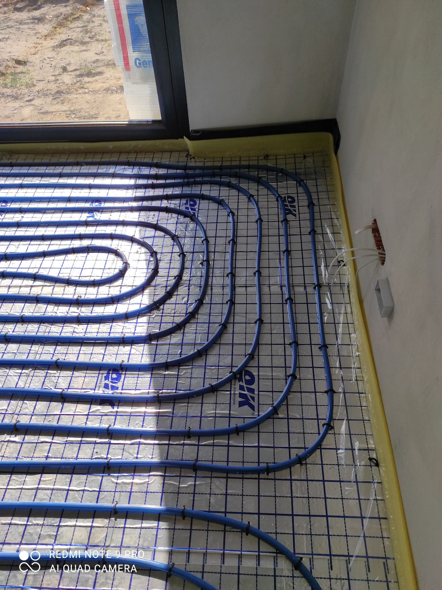 Niebieskie rury ogrzewania podłogowego ułożone na folii z siatką, zabezpieczone czarnymi klipsami. Widoczne kable wystające ze ściany i żółta taśma izolacyjna przy ścianie.