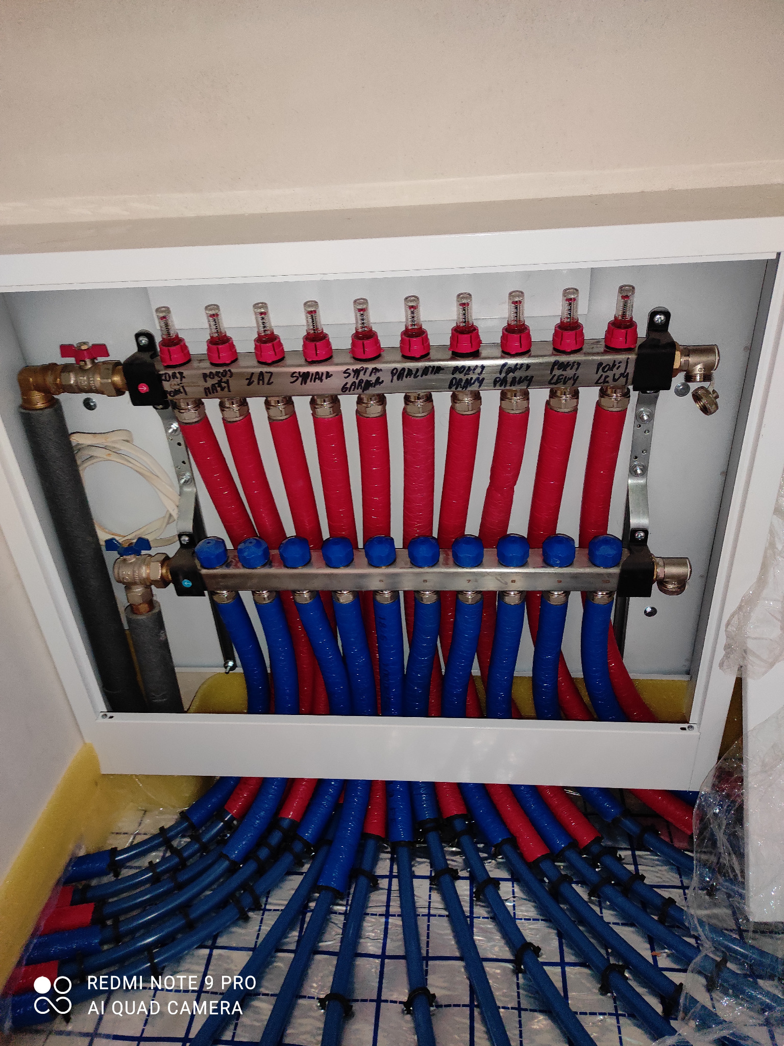 Rozdzielacz ogrzewania podłogowego z rurami w kolorach czerwonym i niebieskim, podpisany markerem, umieszczony w białej szafce instalacyjnej na tle maty izolacyjnej.