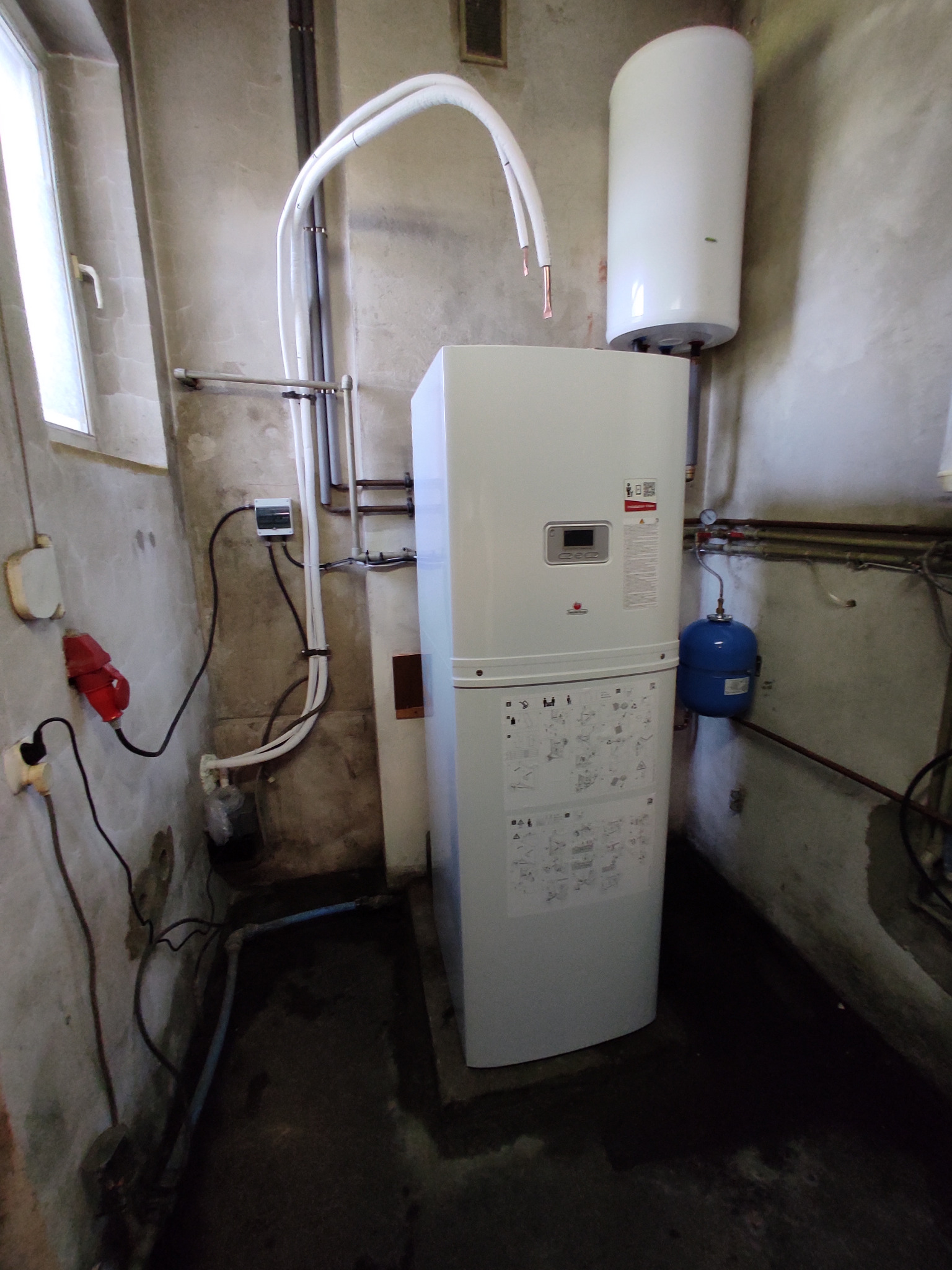 Biała pompa ciepła powietrze-woda z podłączonym zasobnikiem, widoczna w kotłowni z instalacją i osprzętem. Surowe ściany.