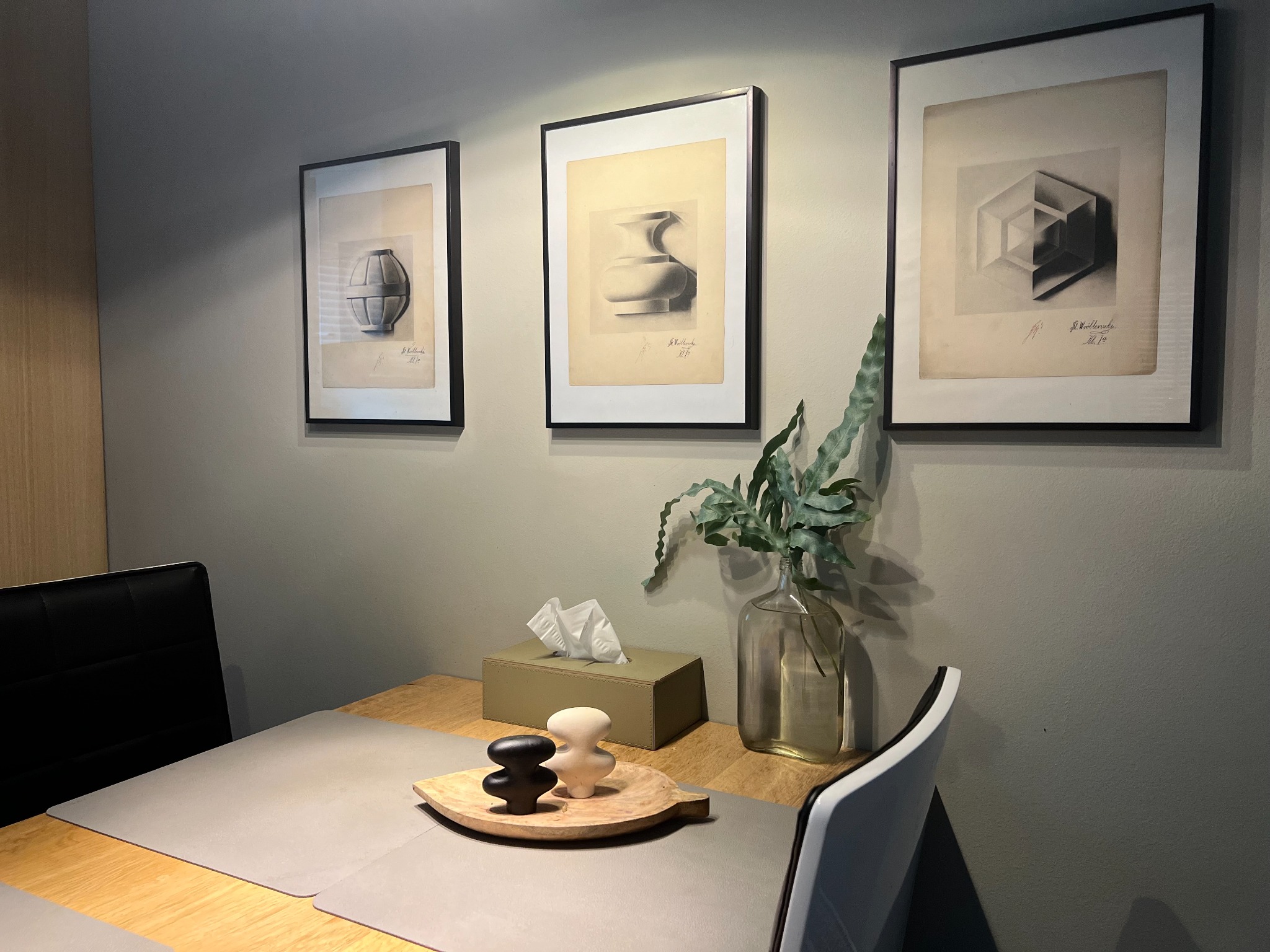 Jadalnia z drewnianym stołem, szarymi podkładkami, dekoracjami i trzema czarno-białymi grafikami w ramach na ścianie. Minimalistyczny wystrój.