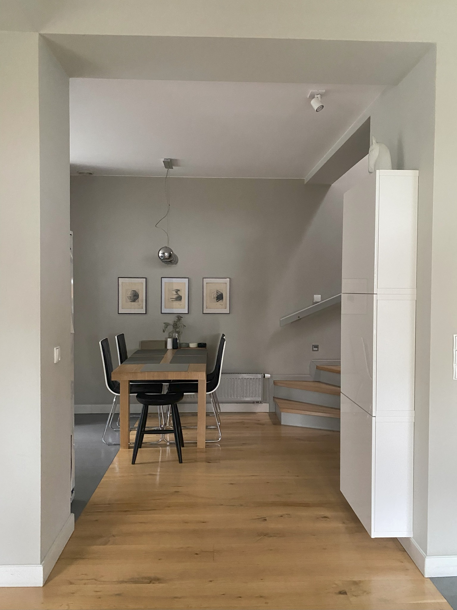 Jasna jadalnia z drewnianym stołem i czarnymi krzesłami, widok z salonu, minimalistyczny design, schody w tle, obrazy na ścianie, podłoga drewniana.