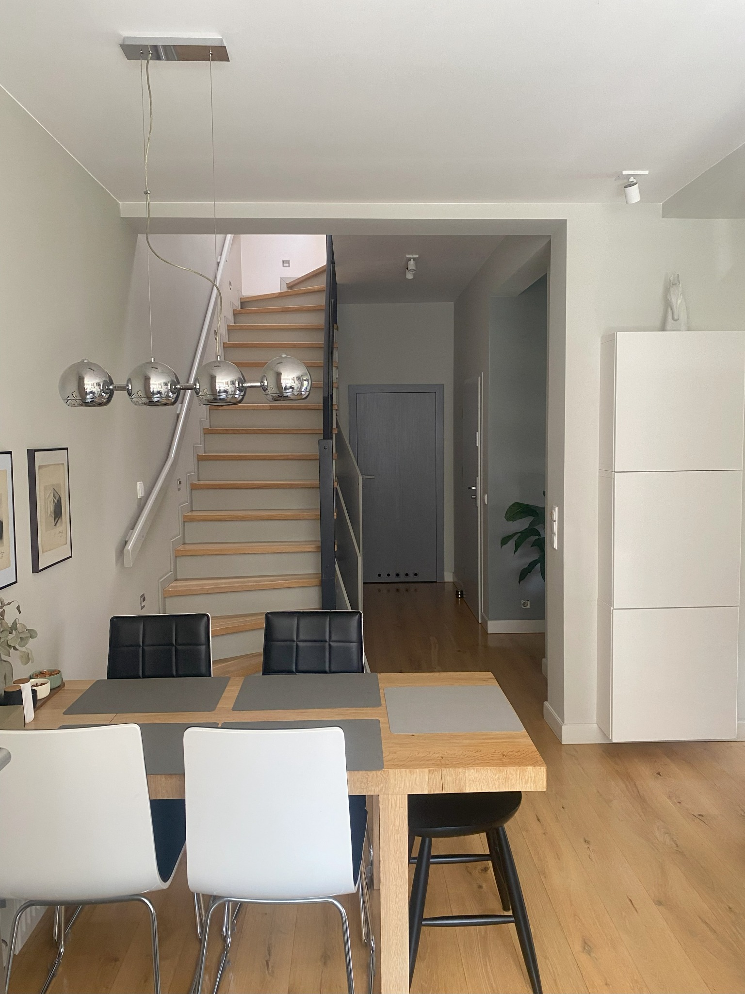 Jasna jadalnia z drewnianym stołem, krzesłami, schodami i srebrną lampą wiszącą. Minimalistyczny wystrój wnętrza z akcentami szarości i bieli, parkiet na podłodze.