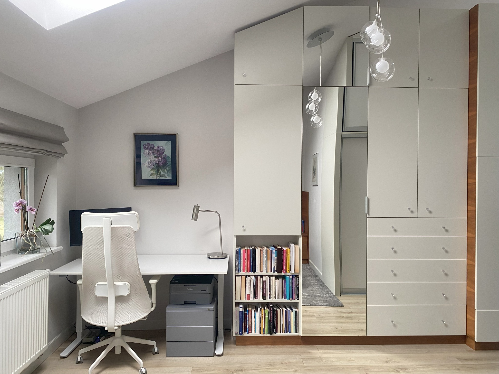 Jasne biuro w domu: białe biurko z komputerem, ergonomiczne krzesło, szafa z lustrem. Skos dachu i okno dodają naturalnego światła. Minimalistyczny design.