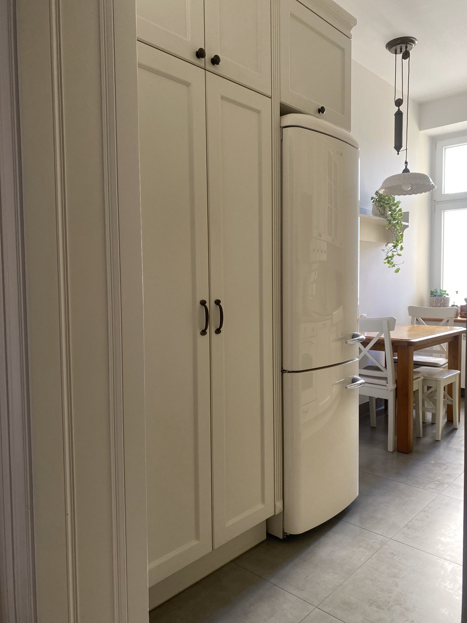 Kuchnia w stylu retro: kremowe szafki z czarnymi uchwytami, lodówka w stylu vintage i drewniany stół z białymi krzesłami, oświetlona wiszącą lampą z obciążnikiem.