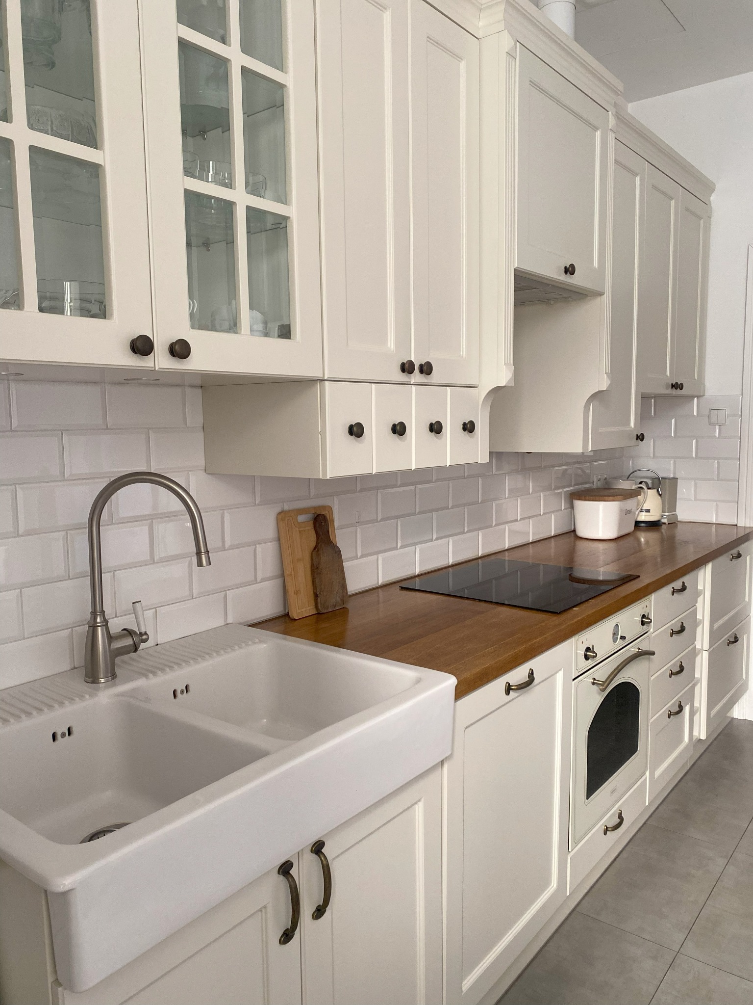 Jasna kuchnia w stylu rustykalnym: białe szafki z ciemnymi uchwytami, drewniany blat, ceramiczny zlew i bateria retro. Uwagę zwraca klasyczna elegancja.