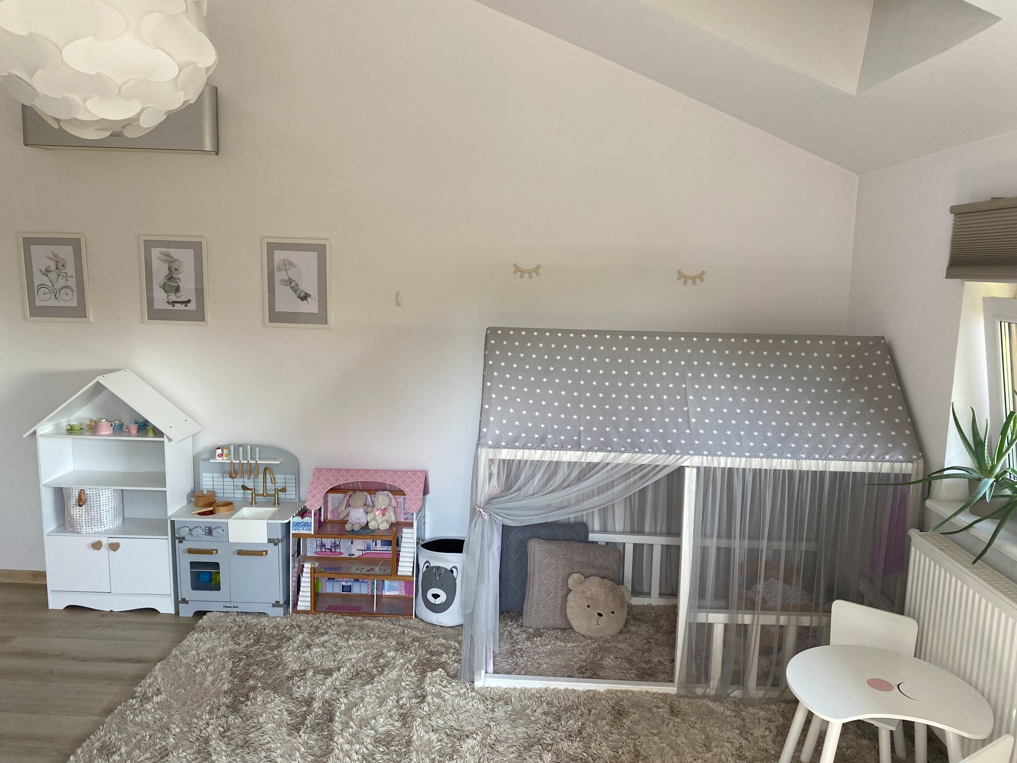 Jasny pokój dziecięcy z domkiem-łóżkiem, drewnianą kuchnią i regałem na zabawki. Uwagę przykuwa puszysty dywan i dekoracyjne naklejki w kształcie rzęs nad łóżkiem.