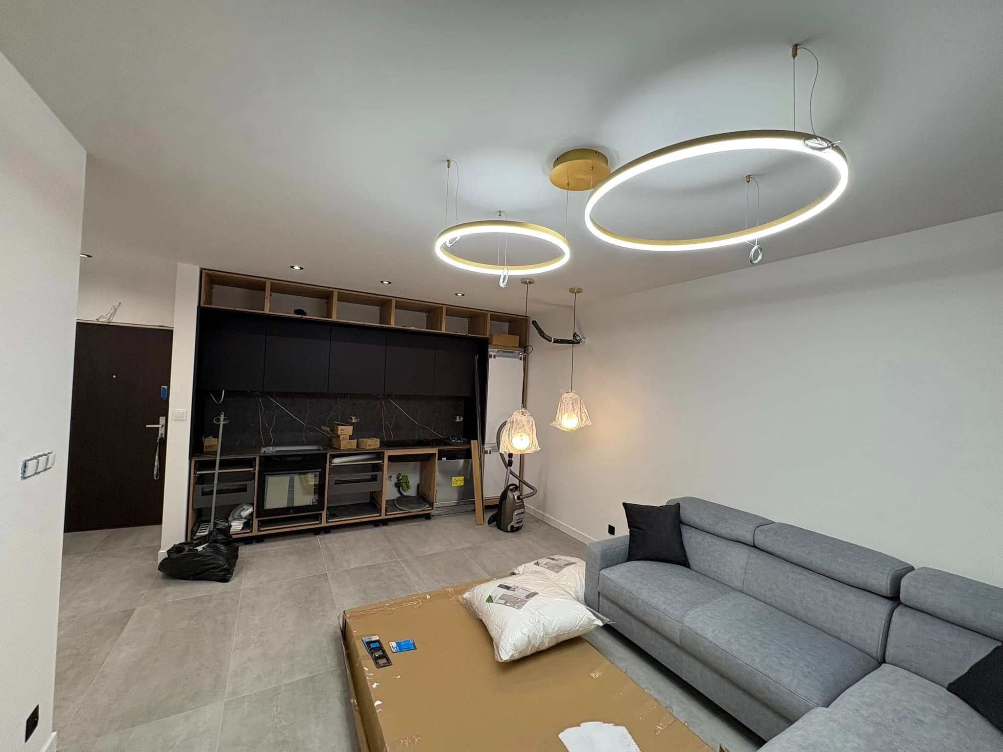 Wnętrze pokoju z szarą sofą, kartonami na podłodze i częściowo zamontowaną zabudową kuchenną z czarnymi frontami i złotymi lampami wiszącymi.