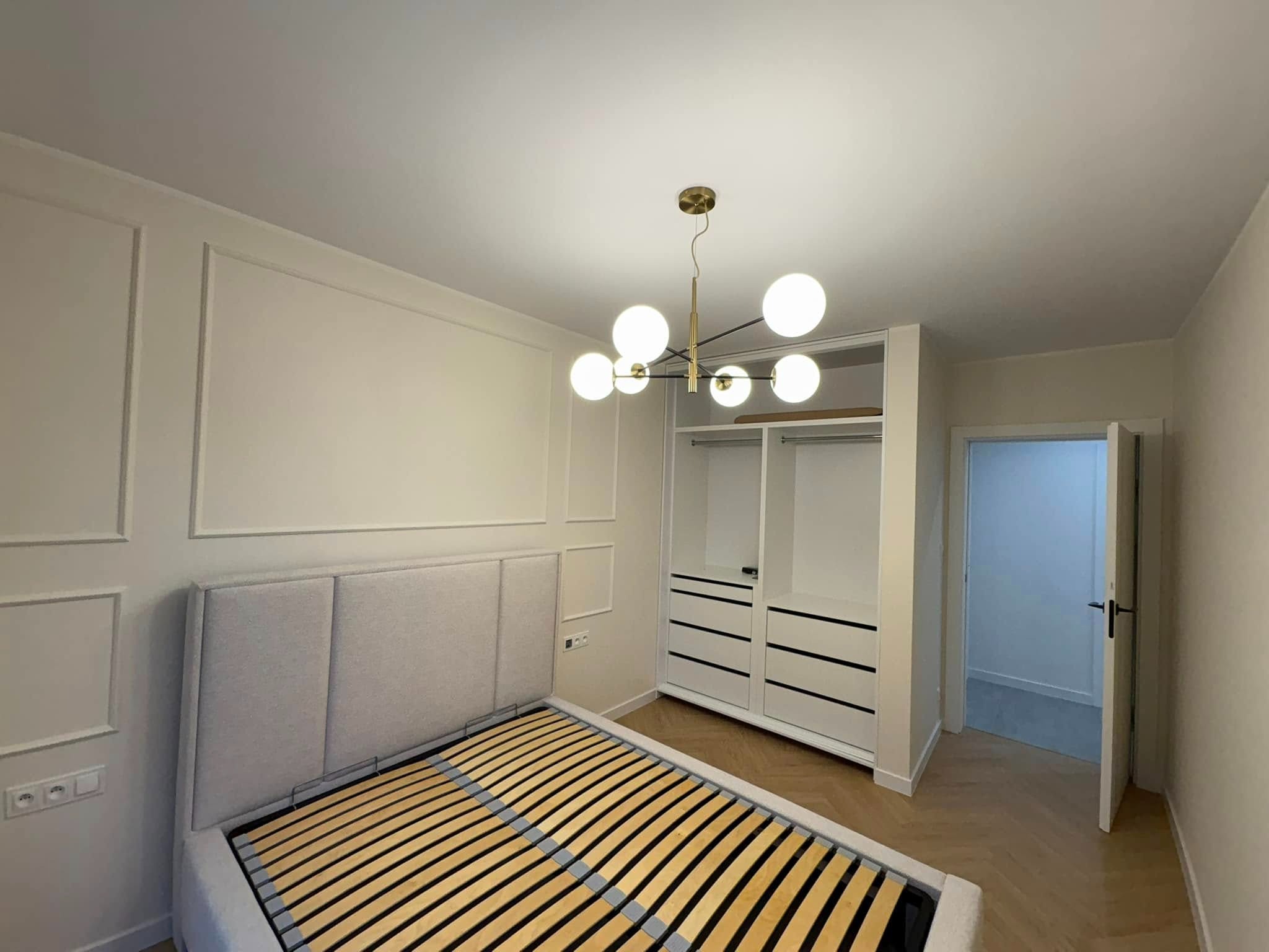 Jasna sypialnia po remoncie: łóżko z szarą ramą, biała szafa wnękowa z czarnymi uchwytami, dekoracyjne listwy na ścianie i parkiet w jodełkę.