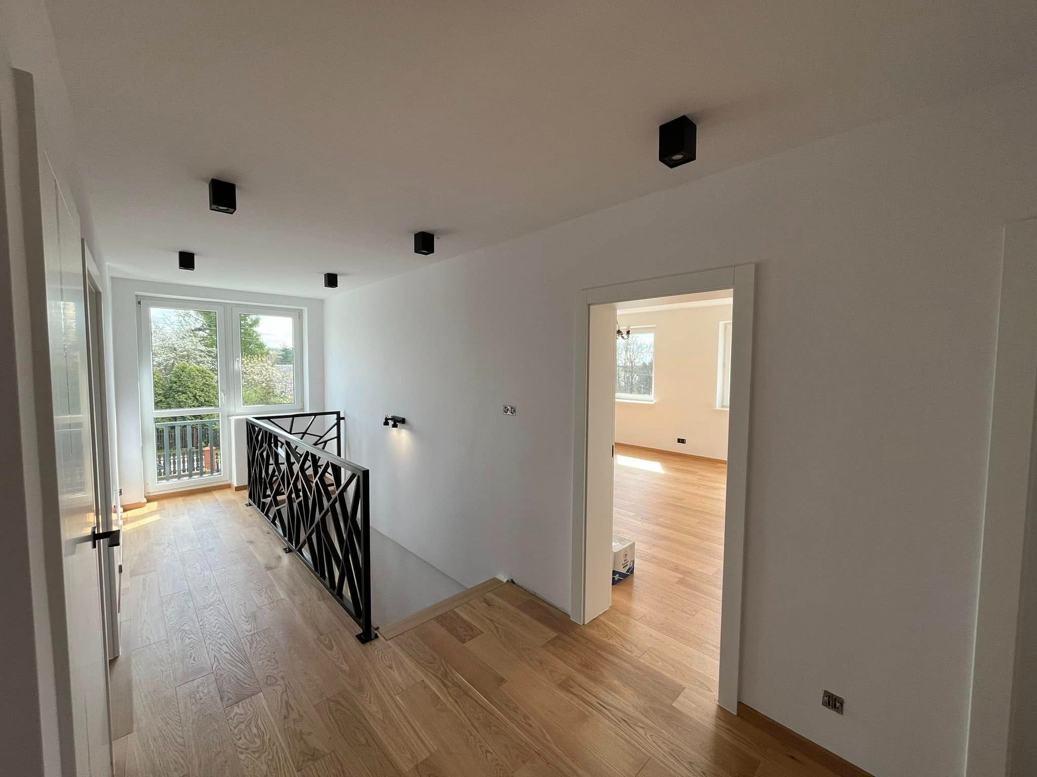 Jasne wnętrze po remoncie: drewniana podłoga, białe ściany, czarne lampy sufitowe i balustrada. Widok z korytarza do pokoju z oknem i zielenią za nim.