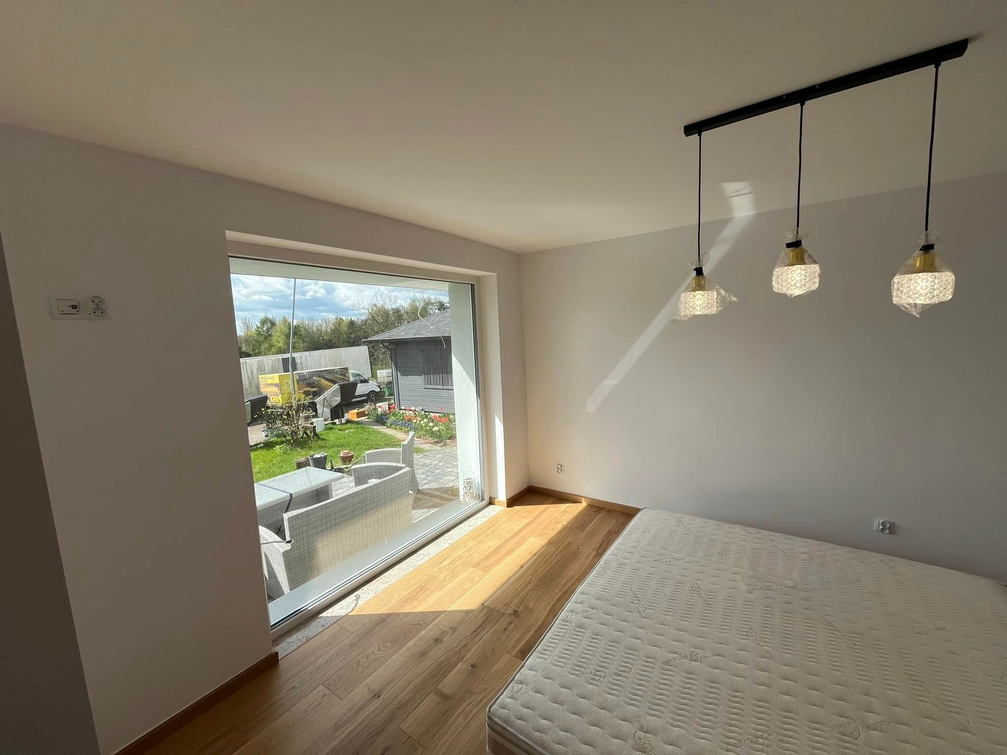 Jasne wnętrze sypialni z parkietem, widokiem na ogród przez duże okno i lampami w foliowych osłonach. Białe ściany, nowy materac.