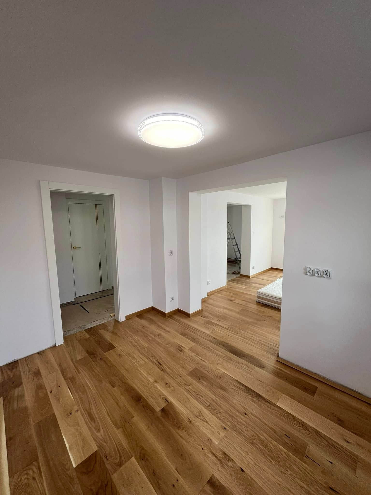 Jasne, odnowione wnętrze z drewnianą podłogą i świeżo pomalowanymi ścianami. Widoczne gniazdka elektryczne i przejścia do innych pomieszczeń. Nowoczesne oświetlenie sufitowe.