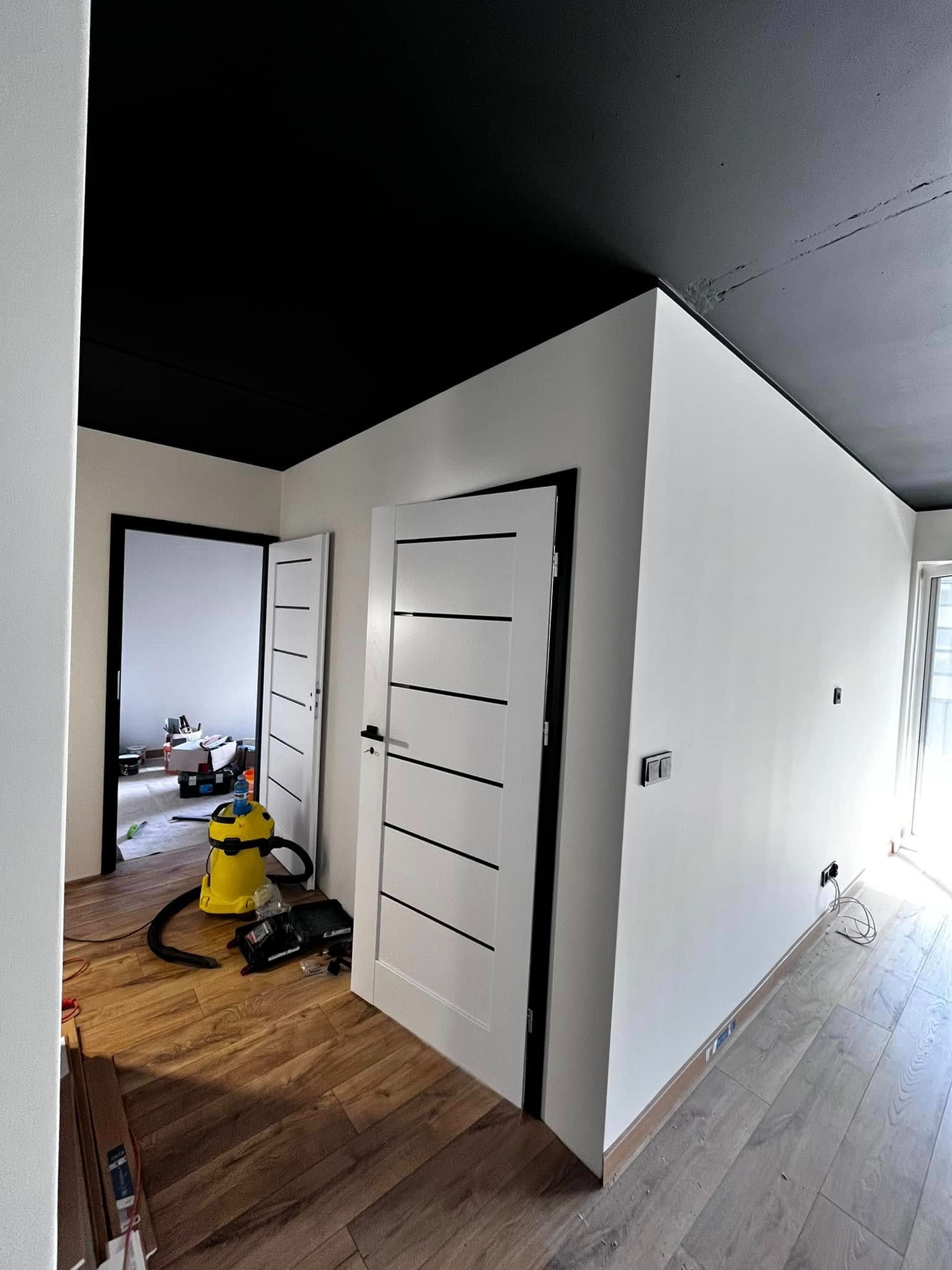 Pokój po remoncie: białe ściany, czarny sufit, drewniana podłoga. Widoczne drzwi wewnętrzne z czarnymi akcentami i sprzęt remontowy, sugerujący trwające prace wykończeniowe.