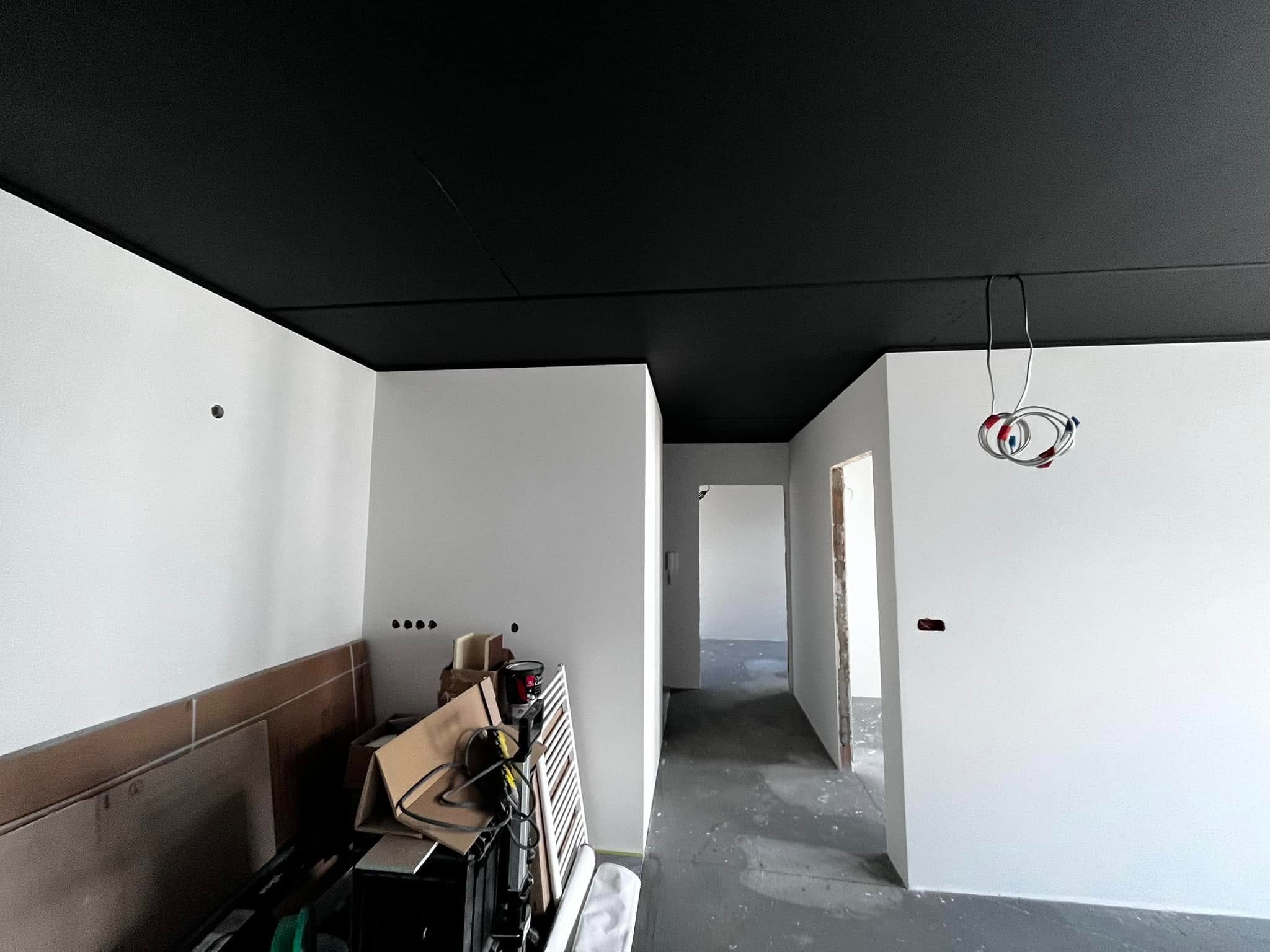 Białe ściany i czarny sufit w wyremontowanym wnętrzu. Widoczne kable, otwory w ścianach i materiały budowlane. Minimalistyczny design w trakcie realizacji.
