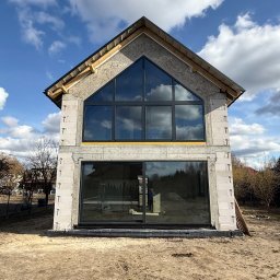 MPS-OKNO DOM KATARZYNA SZKUTNIK - Surowy stan domu z dużymi, czarnymi oknami i drzwiami przesuwnymi, widoczne bloczki i konstrukcja dachu, błękitne niebo z chmurami odbija się w szybach.