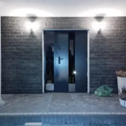 Nowoczesne, grafitowe drzwi wejściowe z przeszkleniami, zamontowane w elewacji z kamienia łupkowego, oświetlone kinkietami. Widoczne schody wejściowe i donice z roślinami.