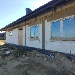 MPS-OKNO DOM KATARZYNA SZKUTNIK - Dom w trakcie budowy z zamontowanymi oknami PCV marki VEKA. Widoczne bloczki, ocieplenie i elementy montażowe. W tle fragment białego busa i sąsiedni budynek.