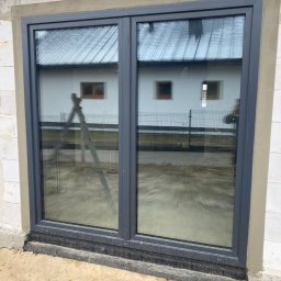 MPS-OKNO DOM KATARZYNA SZKUTNIK - Szare okno PCV dwuskrzydłowe, widoczne z zewnątrz, osadzone w surowym murze. W szybach odbija się biały budynek i drabina oparta o ścianę.