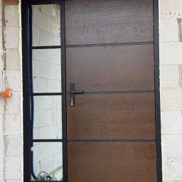 MPS-OKNO DOM KATARZYNA SZKUTNIK - Nowoczesne drzwi zewnętrzne z szybą boczną, brązowe, z czarną ramą, zamontowane w surowym murze w Janowie Lubelskim. Widoczne pianowanie uszczelniające.