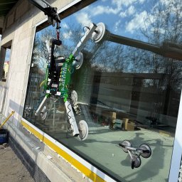 MPS-OKNO DOM KATARZYNA SZKUTNIK - Montaż okna z użyciem przyssawki mechanicznej Glass System DS600 na budowie. W tle widoczne niebo odbijające się w szybie i narzędzia.