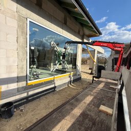 MPS-OKNO DOM KATARZYNA SZKUTNIK - Montaż okna z użyciem przyssawki i żurawia na budowie. Widoczne bloczki betonowe, nieotynkowana ściana i błękitne niebo. Okno duże, z białymi ramami.