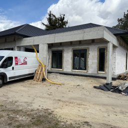 MPS-OKNO DOM KATARZYNA SZKUTNIK - Budynek w stanie surowym z zamontowanymi oknami, na zewnątrz zaparkowany biały samochód dostawczy z logo firmy. Widoczne materiały budowlane i paleta.