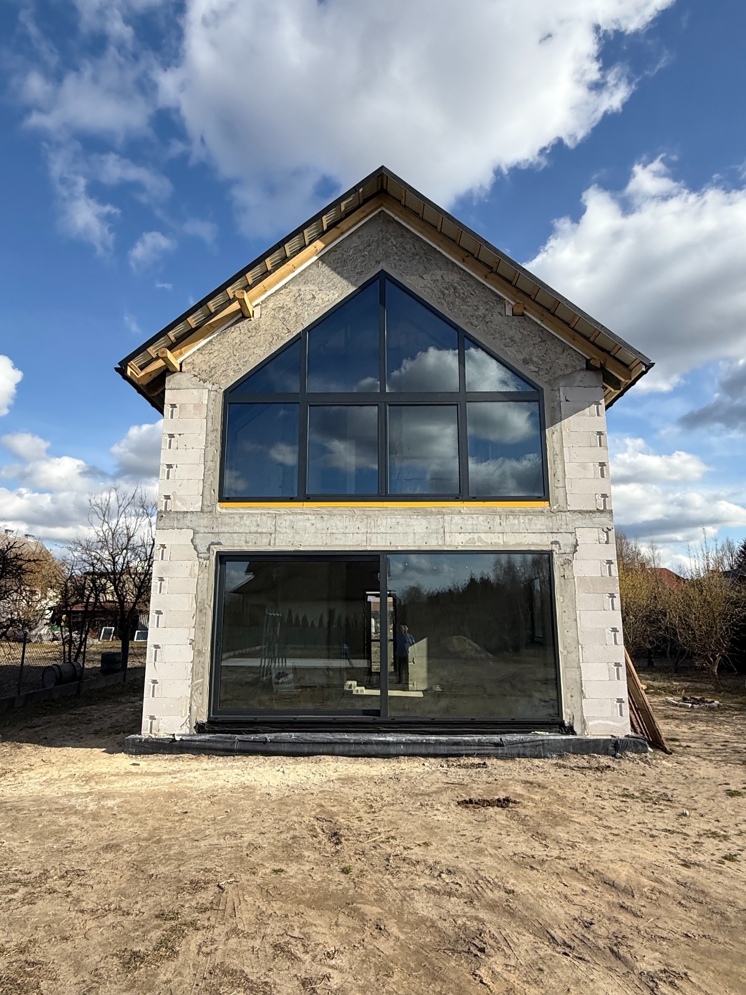 Surowy stan domu z dużymi, czarnymi oknami i drzwiami przesuwnymi, widoczne bloczki i konstrukcja dachu, błękitne niebo z chmurami odbija się w szybach.