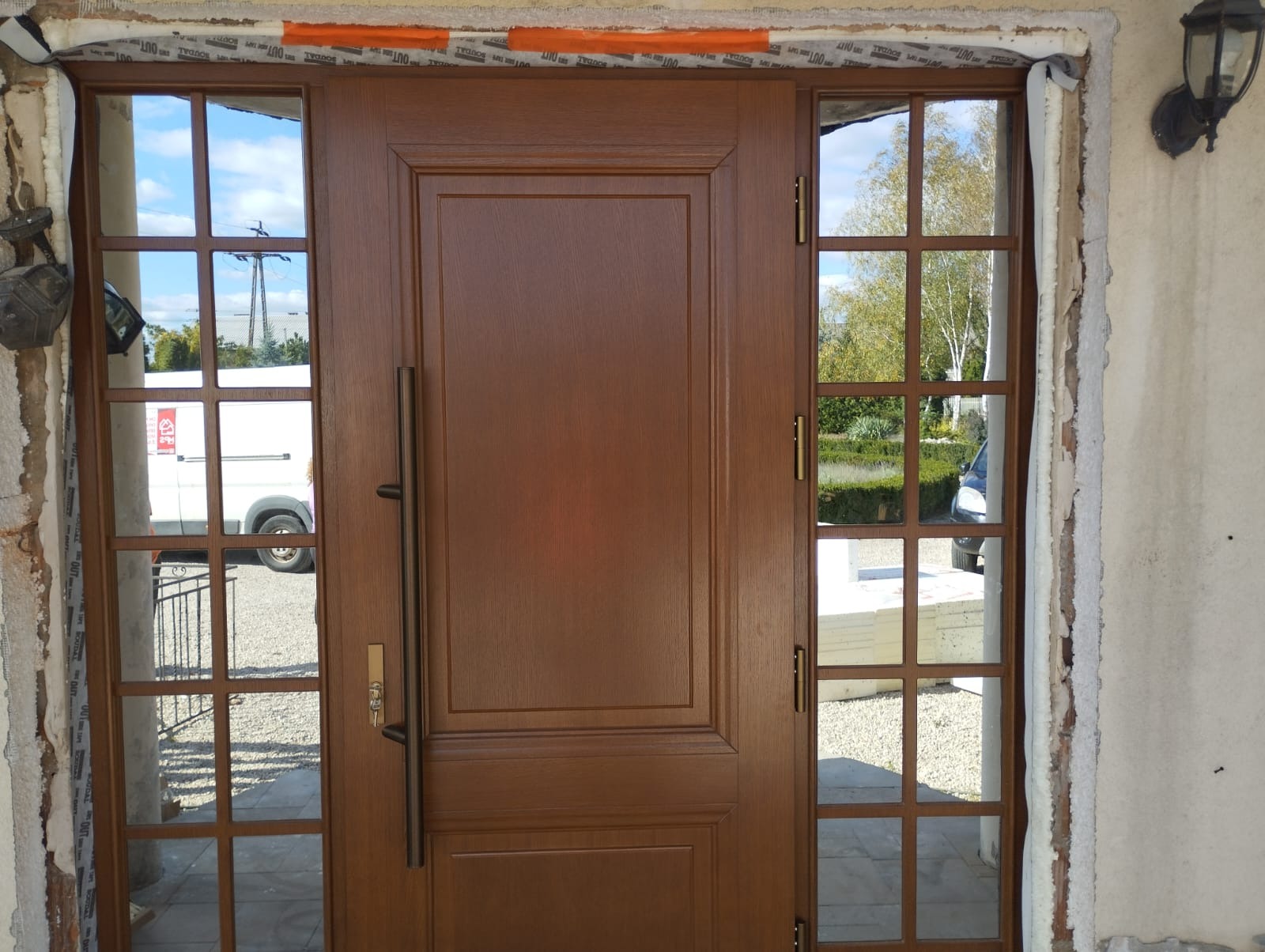 Brązowe drzwi wejściowe z szybami po bokach, zamontowane w surowym otworze, z widocznymi elementami montażowymi i fragmentem elewacji. Długi uchwyt.