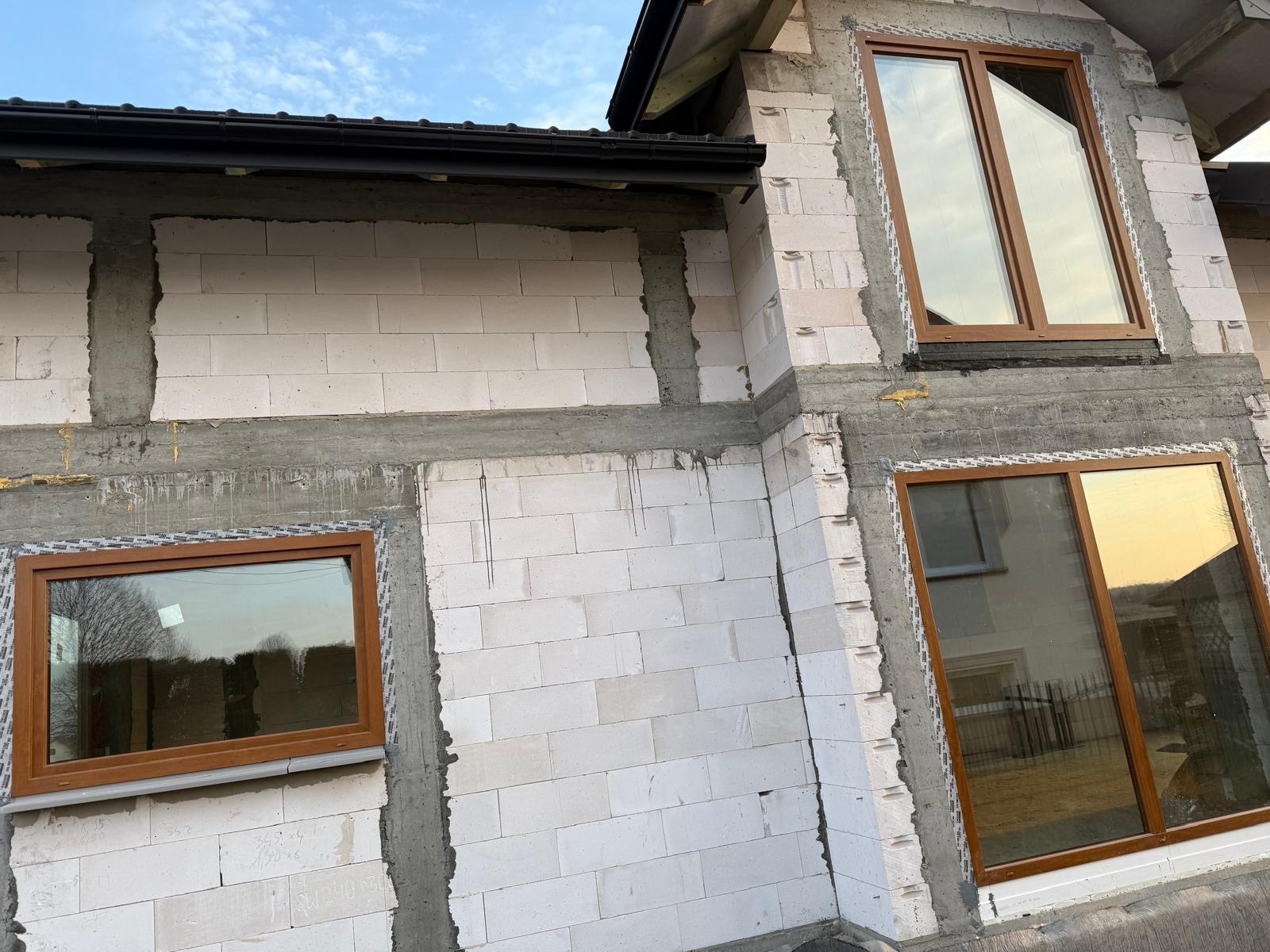 Brązowe okna w surowym budynku z białego bloczka, widoczne ślady montażu, jasne niebo w tle, budowa domu w trakcie prac wykończeniowych.