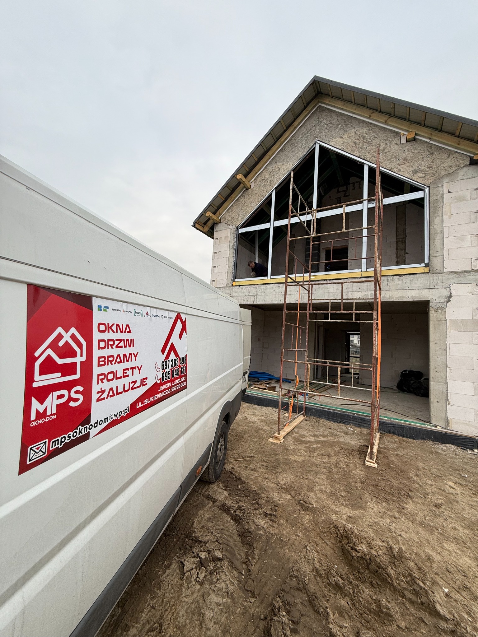 Montaż okien w surowym budynku z rusztowaniem. Na pierwszym planie samochód dostawczy z reklamą firmy MPS Okno-Dom z Janowa Lubelskiego.