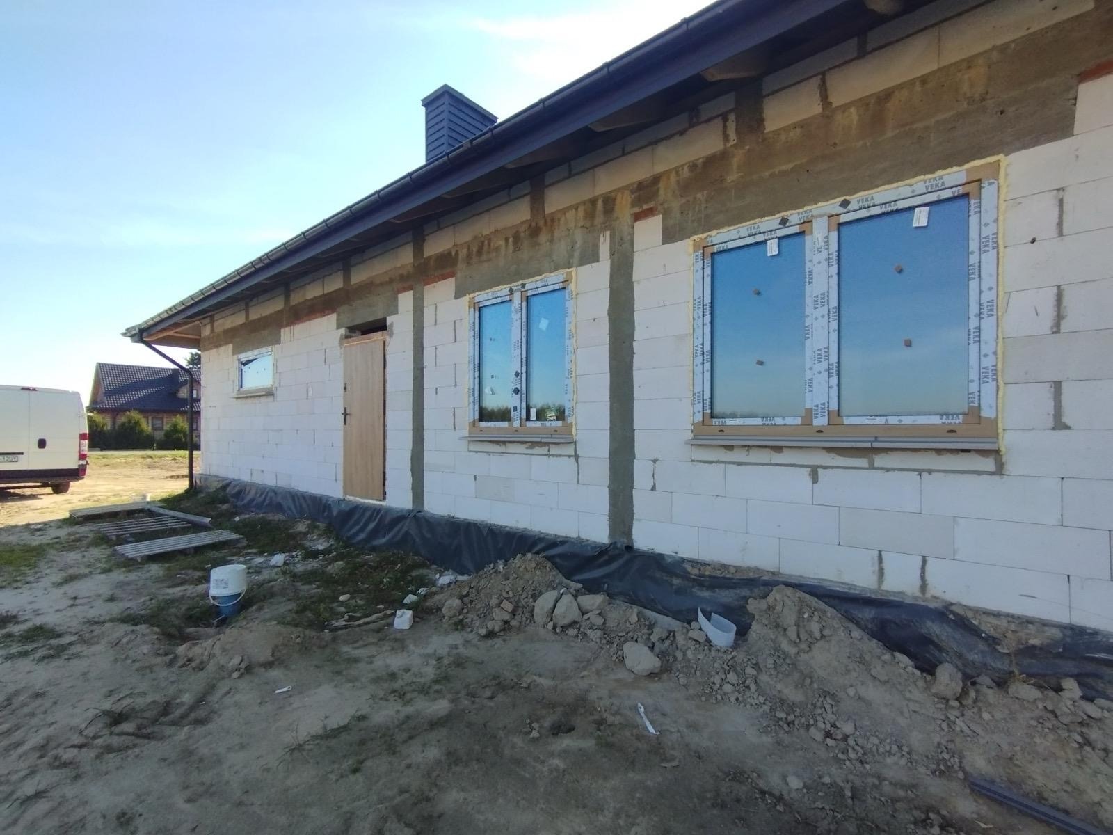 Dom w trakcie budowy z zamontowanymi oknami PCV marki VEKA. Widoczne bloczki, ocieplenie i elementy montażowe. W tle fragment białego busa i sąsiedni budynek.