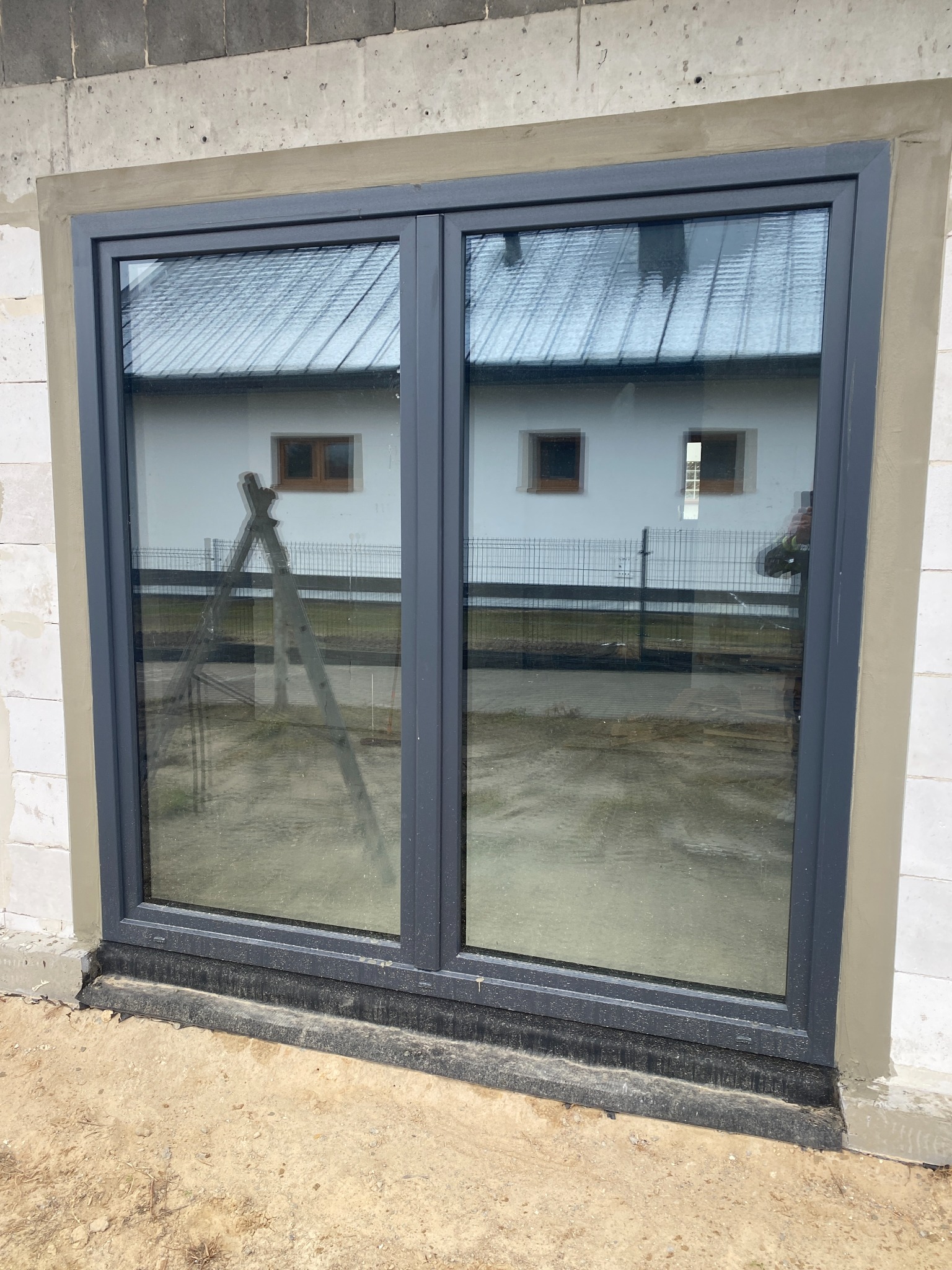 Szare okno PCV dwuskrzydłowe, widoczne z zewnątrz, osadzone w surowym murze. W szybach odbija się biały budynek i drabina oparta o ścianę.